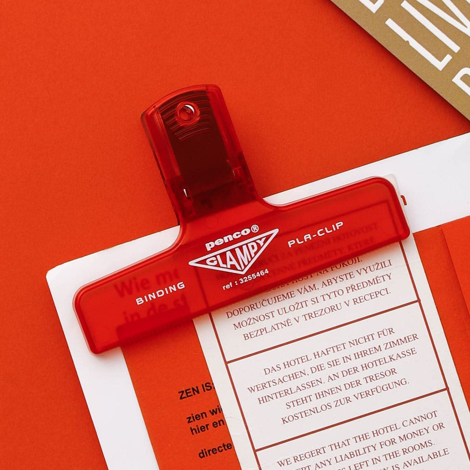 A red Hightide Penco Clampy Plastic Clip securing a sheet of paper on a vibrant orange clipboard.