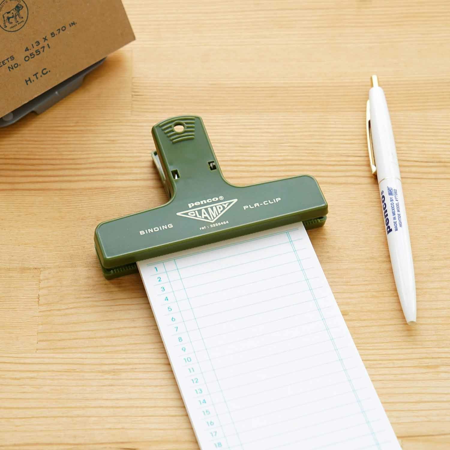 A green Hightide Penco Clampy Plastic Clip holding a notepad in place on a wooden desk beside a white pen.
