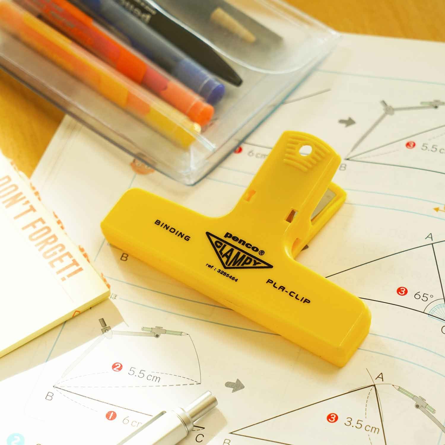 A yellow Hightide Penco Clampy Plastic Clip placed on a workspace with pens, scattered paper, and stationery.
