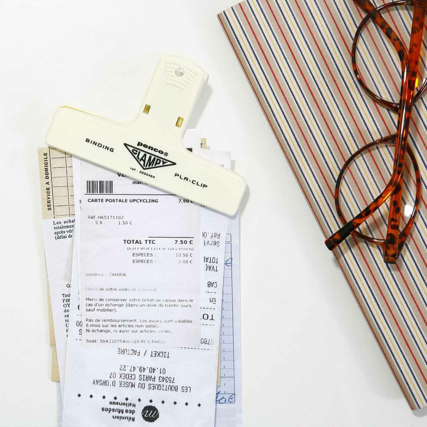 A cream Hightide Penco Clampy Plastic Clip holding documents on a striped surface with red glasses nearby.