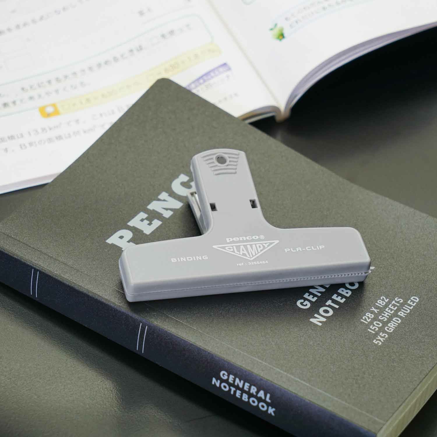 A grey Hightide Penco Clampy Plastic Clip resting on an olive green notebook with an open planner beside it.
