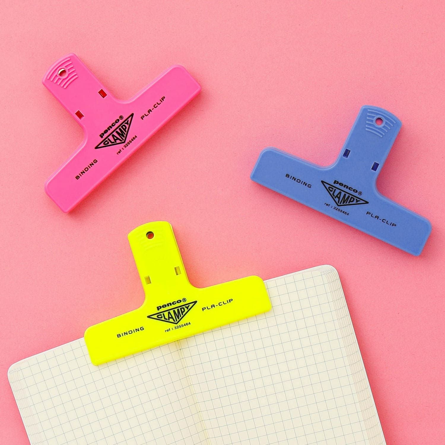Three Hightide Penco Clampy Plastic Clips in pink, neon yellow, and blue holding lined papers on a pink background.