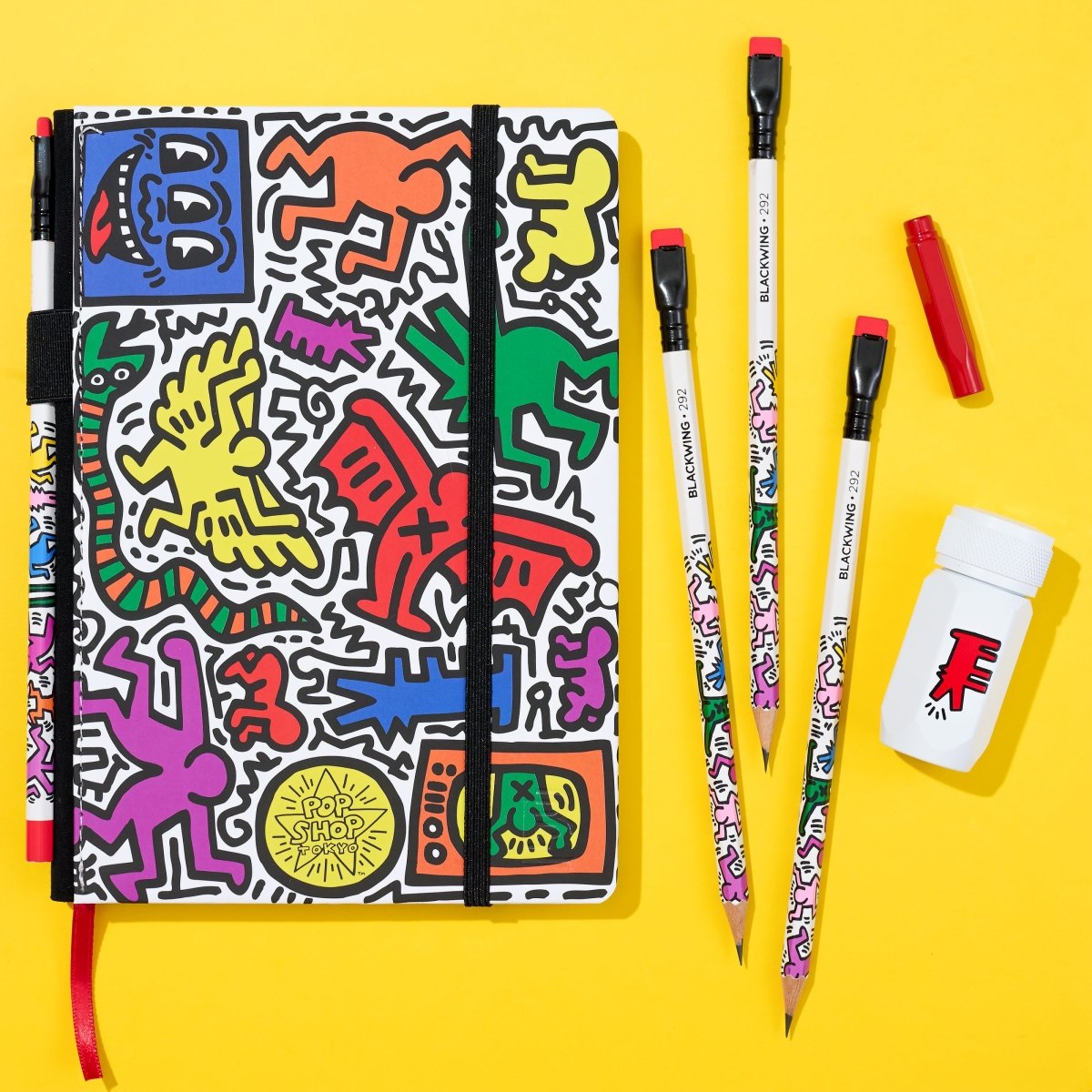 Flat lay of the set on a yellow background, displaying the sketchbook with Keith Haring artwork and included writing tools.