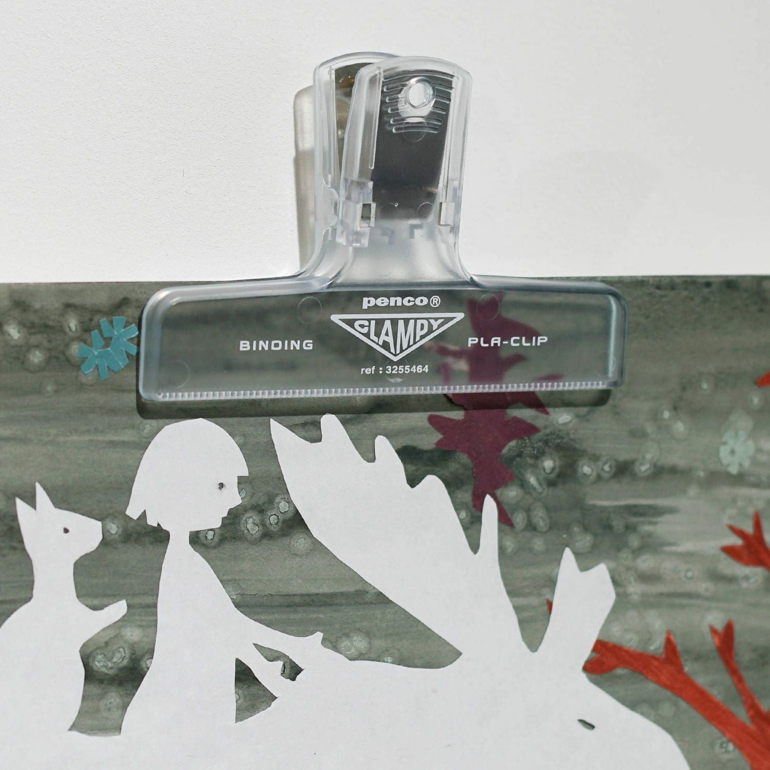 A close-up of a clear Hightide Penco Clampy Plastic Clip holding a paper cut-out against a grey background.
