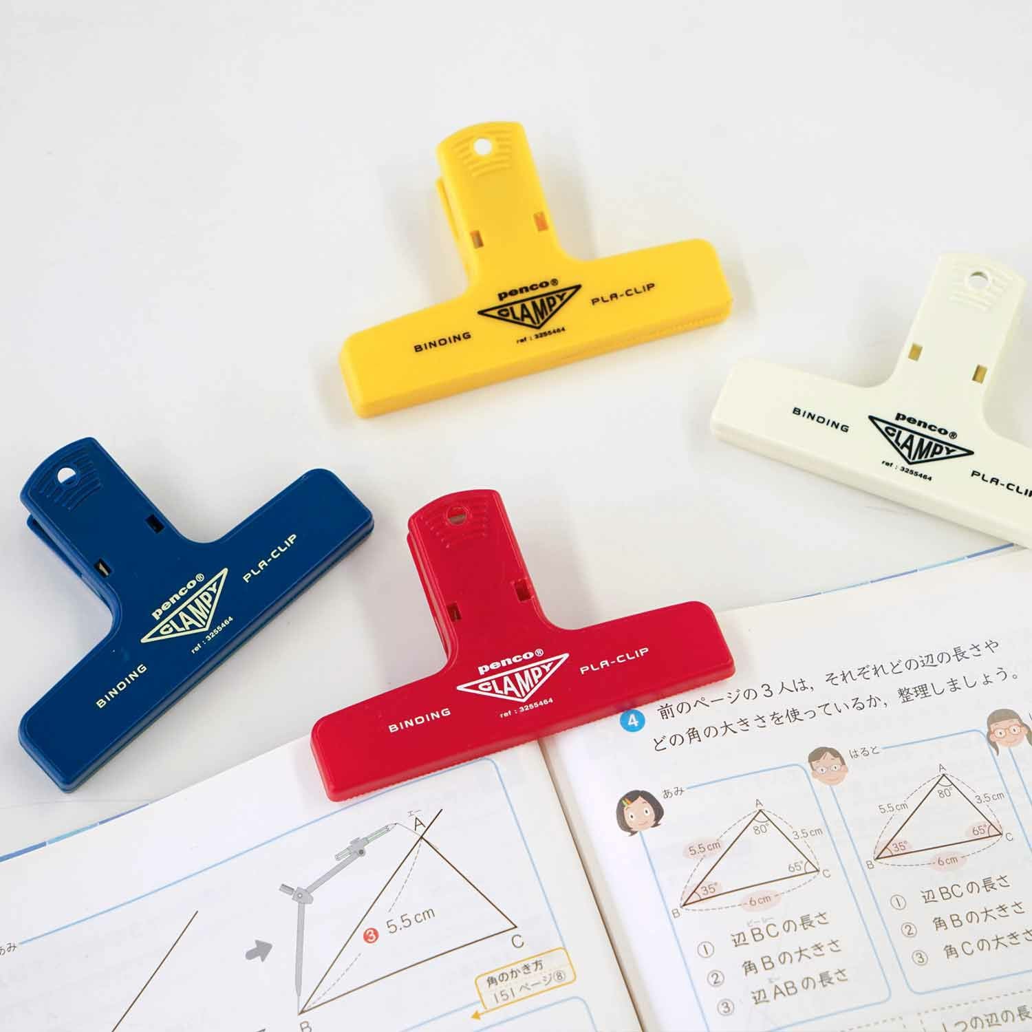 Three Hightide Penco Clampy Plastic Clips in yellow, blue, and red holding various sheets of paper with printed Japanese notes.