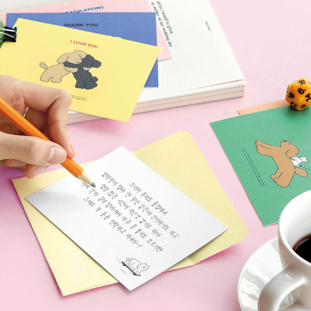 Person writing on Iconic Cuddly Message Card beside open yellow box and stationery items.