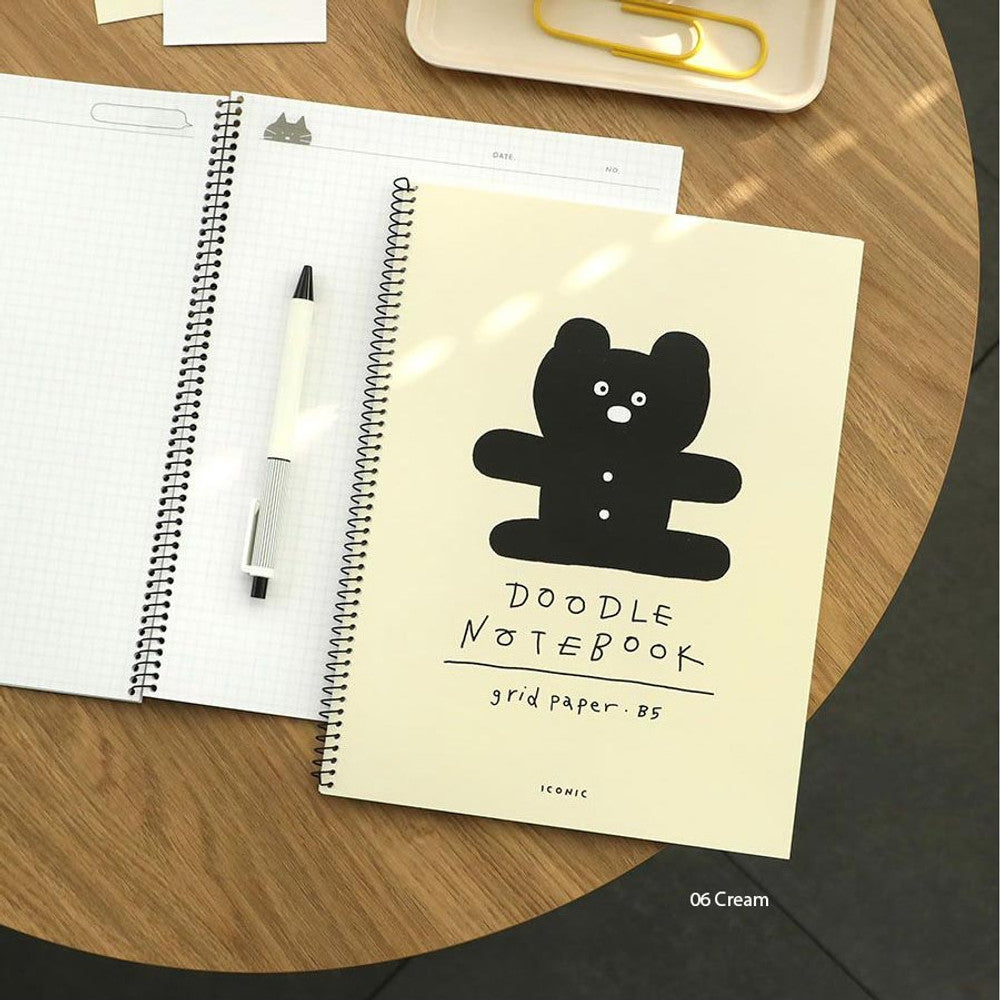 Iconic Doodle B5 Grid Notebook in beige and black covers displayed on wooden desk with pen.