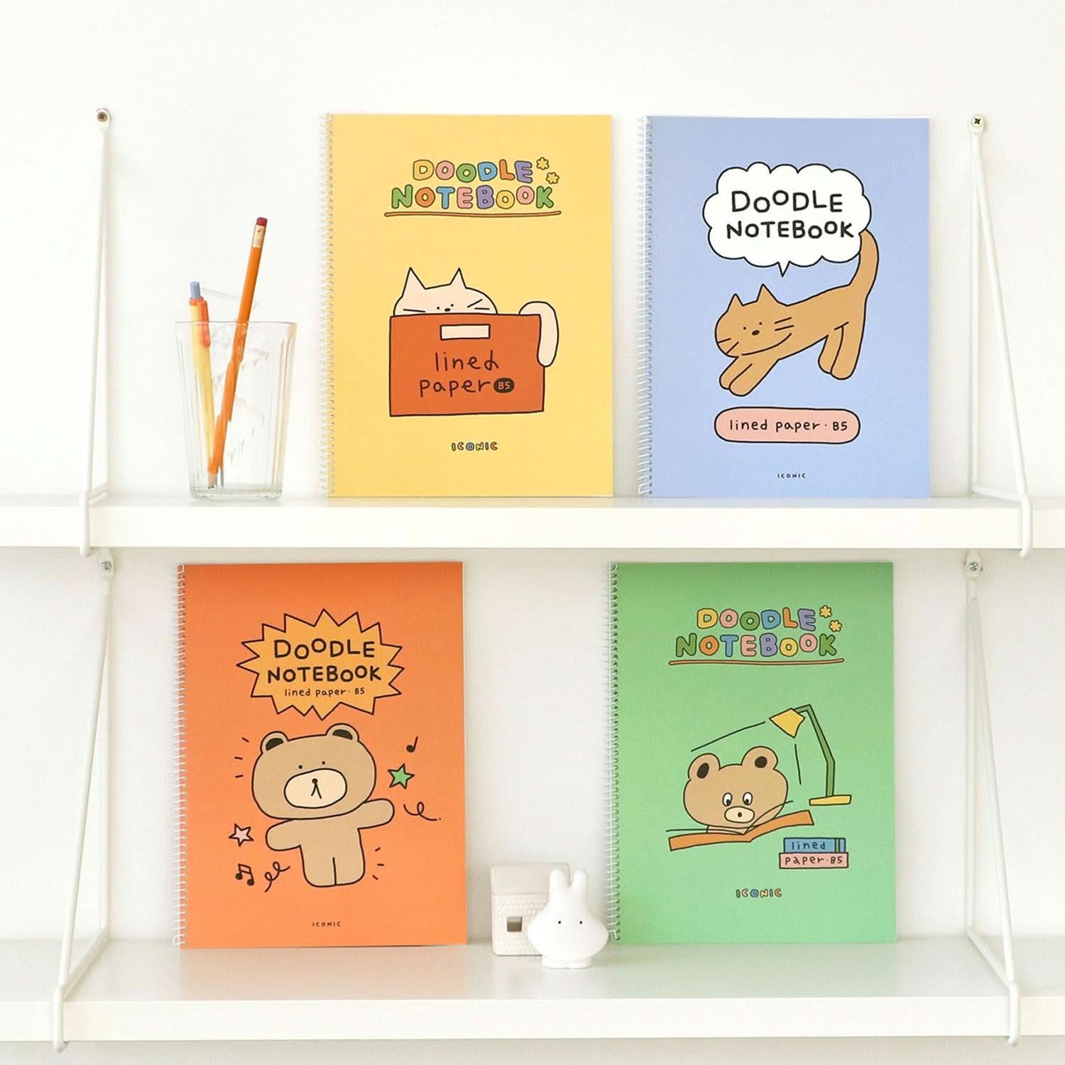 Iconic Doodle B5 Spiral Notebooks in orange, green, blue, and yellow with playful illustrated covers displayed on white shelf.