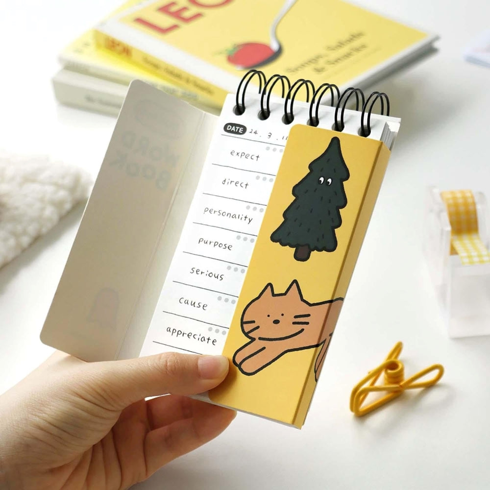 A person holding a compact yellow Iconic Doodle Word Pocket Size Notebook with a cute tree and cat design on the cover.