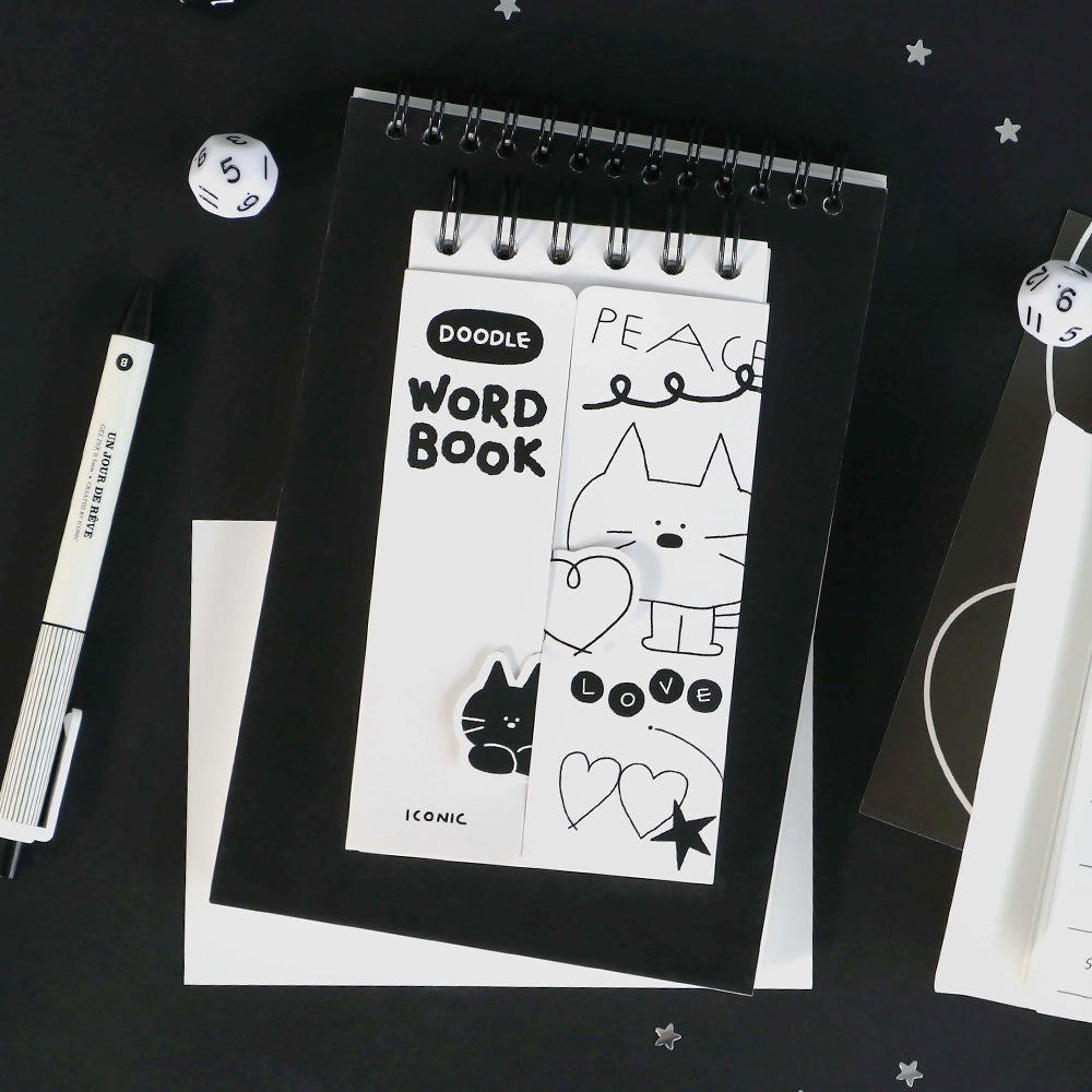 A white Iconic Doodle Word Compact  Size Notebook with playful doodle-style characters, shown on a dark workspace beside a pen.