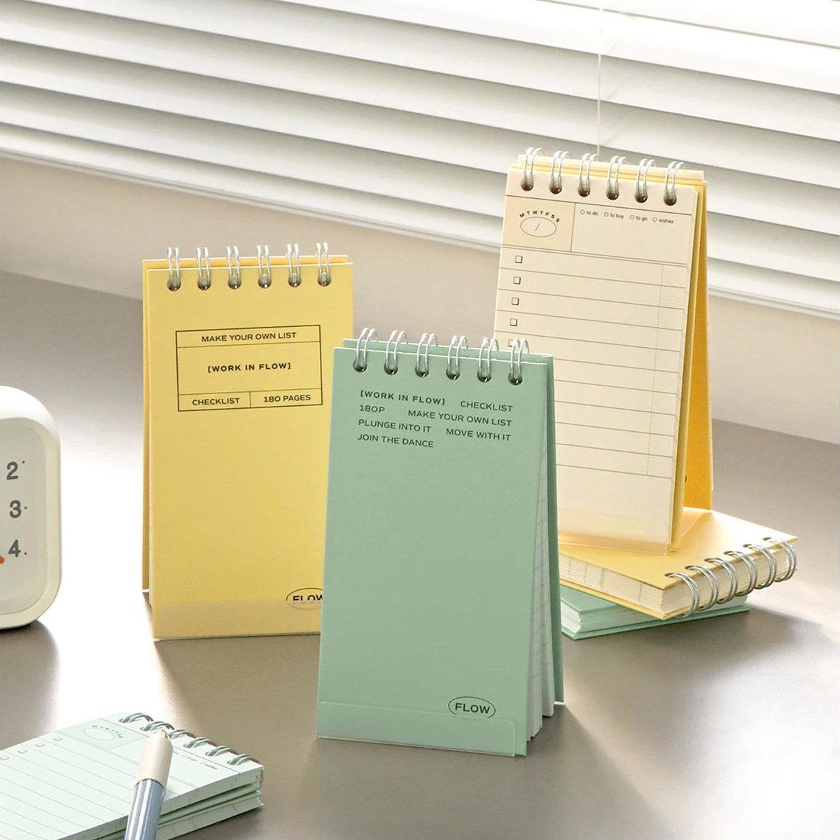 Two Iconic Flow Checklists in yellow and mint green standing on a bright desk by a window, with light streaming through the blinds.