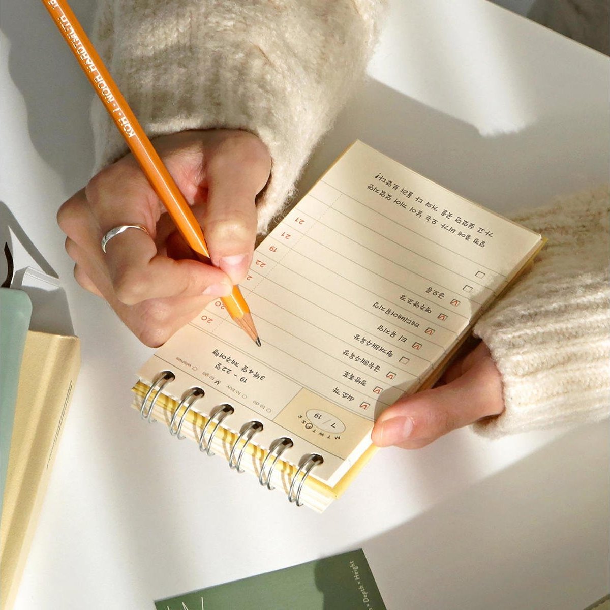 A person writing in the yellow Iconic Flow Checklist while resting it on a desk, with a pencil in hand.