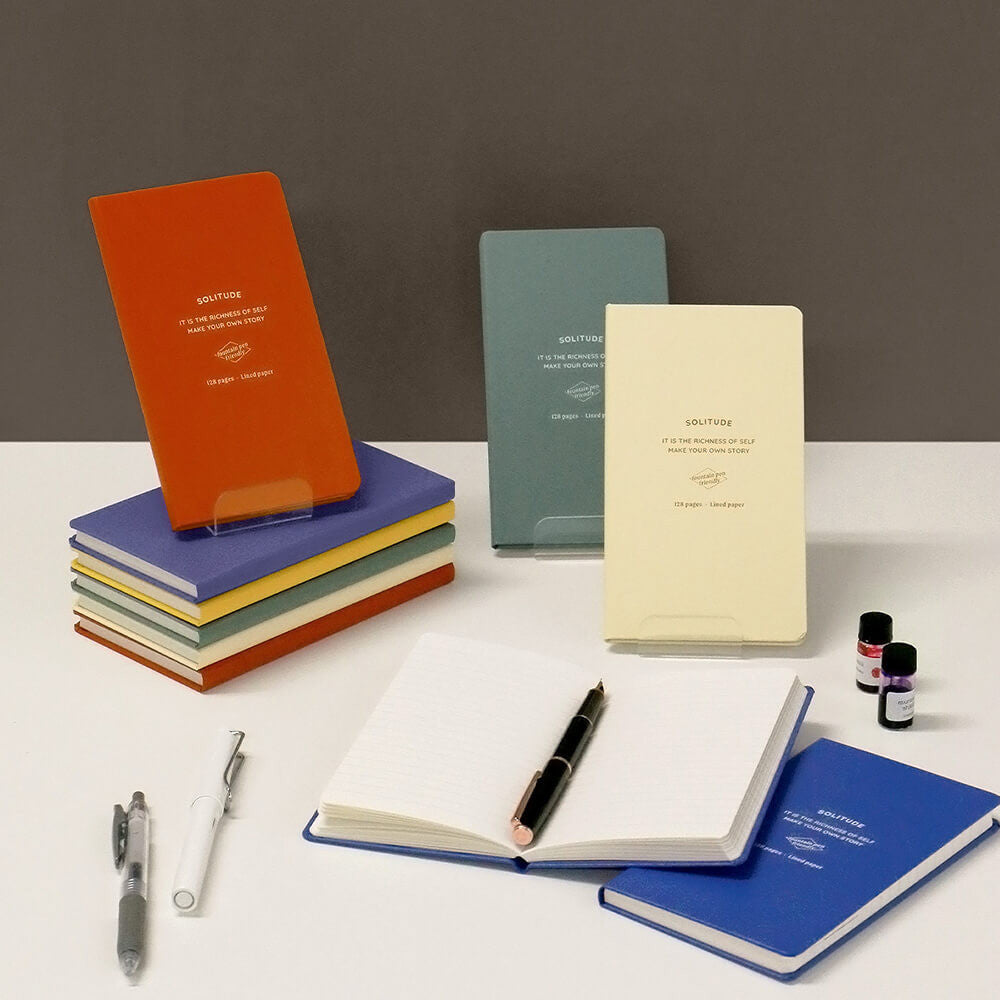 Iconic Solitude B7 Handy Notebooks in assorted colours stacked neatly on desk with pen and ink bottle.