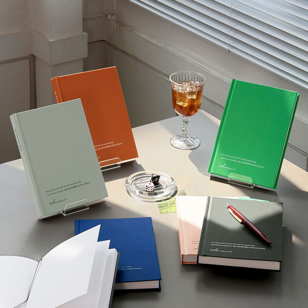 A collection of Iconic Souvenir B6 Grid Notebooks in orange, green, blue, and black displayed on a bright desk.