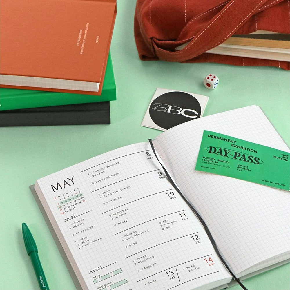 An orange Iconic Souvenir B6 Grid Notebook beside an open notebook with schedule and pen.