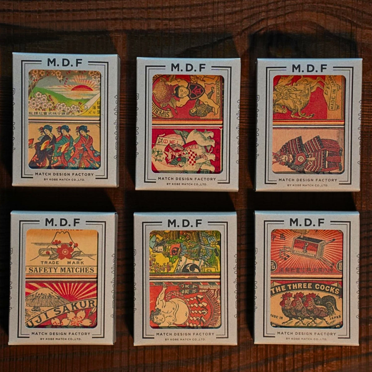 Six M.D.F. Match Design Factory matchboxes with various designs on a wooden surface.